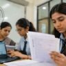 CBSE News Today को दर्शाती हाई-क्वालिटी इमेज जिसमें छात्राएं CBSE बोर्ड की आंसरशीट चेक करती हुई दिखाई दे रही हैं और री-इवैल्यूएशन प्रक्रिया का माहौल नजर आ रहा है।