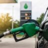 Petrol pump par car me fuel bharte hue green nozzle ka close-up shot, background me fuel prices display board dikh raha hai.